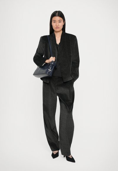 Veste de costume noire texturée et pantalon large, associés à un haut noir et des escarpins noirs à bout pointu. Tenant un sac à main en cuir bleu marine.