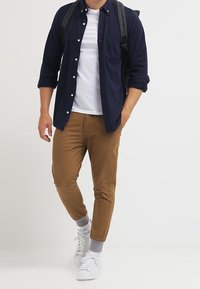 Camicia navy con bottoni sopra una t-shirt bianca, pantaloni beige rastremati, calzini grigi risvoltati e sneakers bianche. Spalline dello zaino visibili sulle spalle.
