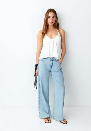 Jeune femme debout contre un mur blanc, portant un haut blanc sans manches, un jean bleu clair à jambes larges, des tongs, et tenant une écharpe noire dans la main gauche.