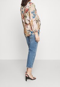 Bloes met bloemenmotief in beige, met kleurrijke patronen, lange mouwen en een ontspannen pasvorm, gecombineerd met lichtblauwe rechttoe-rechtaan jeans en zwarte sandalen.