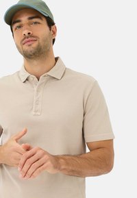 Beige katoenen polo met een twee-knopssluiting en korte mouwen, gecombineerd met een lichtgroen pet. Handen voor zich neerzijds.