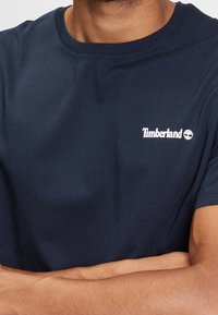 Man klädd i en marinblå Timberland-t-shirt med vitt logotyp på bröstet, med korsade armar, mot en enkel bakgrund.