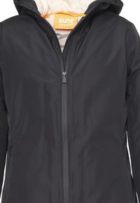 Giacca nera impermeabile con chiusura frontale a zip, cappuccio e morbida fodera beige. Presenta un'etichetta gialla con il logo "SUNS BOARDS" all'interno.