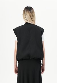 Persoon met lang blond haar draagt een zwart mouwloos gewatteerd vest over een wit T-shirt met korte mouwen en een zwarte rok, met de rug naar de camera gekeerd op een witte achtergrond.