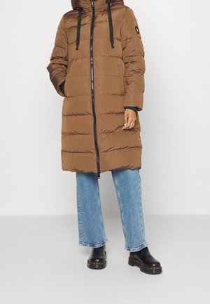 Personne portant un long manteau matelassé marron avec une fermeture éclair à l’avant, un jean bleu et des bottines noires, debout devant un fond uni.
