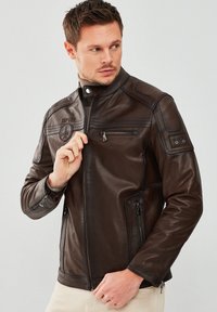 Brown leather jacket with a zippered front and side pockets, featuring shoulder accents and a small logo on the chest. Smooth texture.
