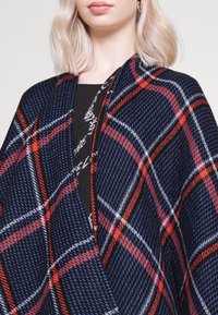 Femme portant un châle à carreaux bleu, rouge, blanc et noir par-dessus un haut noir, avec des cheveux blonds courts et des boucles d'oreilles créoles dorées.