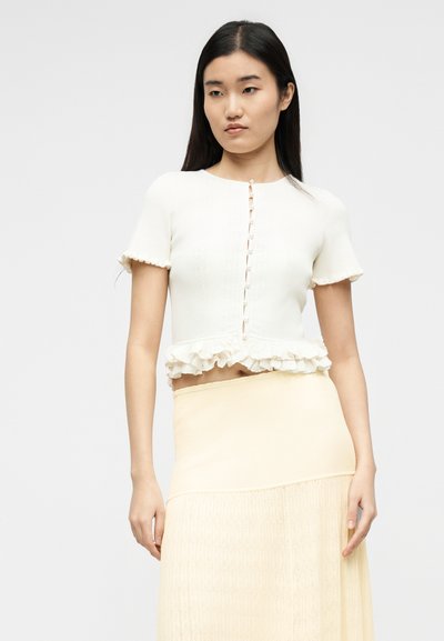 Γυναίκα που φοράει λευκό κοντομάνικο πουκάμισο με κουμπιά και βολάν στο τελείωμα και μια κρεμ πλεκτή φούστα με ψηλή μέση, πάνω σε απλό φόντο.