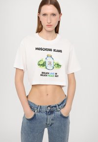 Balts īsās piedurknes t-krekls ar tekstu "MOSCHINO JEANS" un grafisku trauku zaļos akcentos. Kombinēts ar gaiši ziliem, nodilušiem džinsiem.
