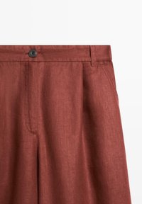 Pantalon en lin couleur rouille avec fermeture par bouton, poches latérales et passants de ceinture visibles à la taille.