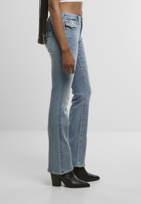 Moteris, vilkinti išblukusias mėlynas bootcut džinsus ir juodas aukštakulnes aulinukus, stovi šonu prieš paprastą baltą foną.