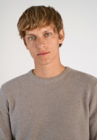 Suéter de punto texturizado gris con cuello redondo, mostrado en una persona con cabello corto castaño claro y ojos azul claros sobre un fondo blanco.