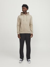 Beige hoodie with a front pocket, adjustable drawstring hood, and ribbed cuffs, paired with black trousers and black sneakers.