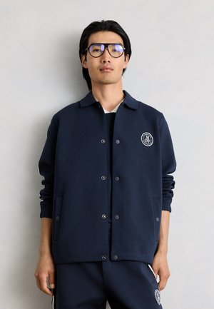 Noor mees mustade prillidega kannab tumesinist jopet ja sobivaid pükse valge embleemiga, seisab lihtsa helehall tausta ees.