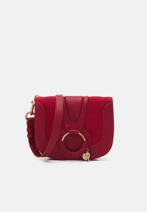 Pequeño bolso bandolera de cuero rojo con anillo dorado y detalle trenzado en el frente, correa ajustable y pequeño adorno dorado colgando del anillo.