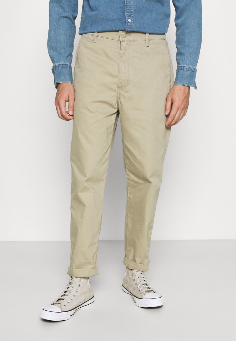 Homme portant un pantalon chino beige aux ourlets retroussés, des baskets montantes gris clair et une chemise en denim bleu boutonnée.