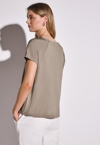 Lys olivengrøn kortærmet bluse med en afslappet pasform og rund kant, lavet af blødt stof. Vist med hvide bukser.