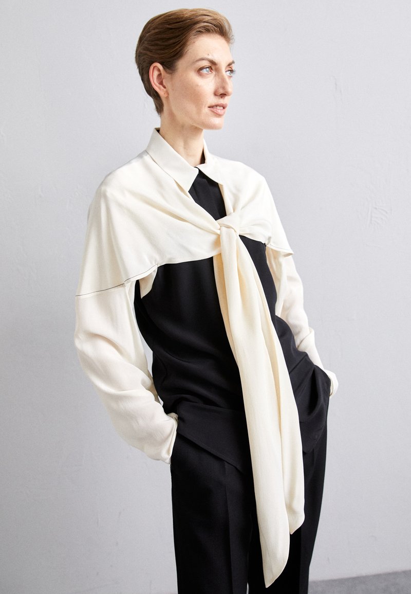 Černobílý outfit s černou blůzou s detailem vázání, pod kterou je vrstvený krémový šál. Elegantní design se strukturovanými tvary.