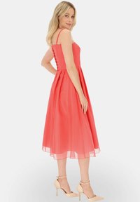 Koraliowa sukienka w kształcie litery A z cienkimi ramiączkami, detalem z sznurowaniem z tyłu oraz półprzezroczystą spódnicą. Noszona z cielistymi szpilkami.