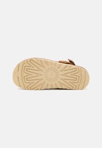 Semelle d'une sandale beige avec des motifs géométriques profonds assurant la traction et un petit logo circulaire près du talon, présentée sur un fond uni.