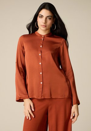 Kobieta o długich ciemnych włosach, nosząca duże koła kolczyków oraz rdzawy, zapinany na guziki długi rękaw i dopasowane spodnie na beżowym tle.