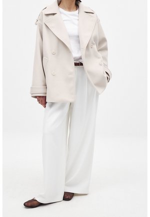Personne portant un manteau beige croisé sur une chemise blanche et un pantalon blanc large avec des chaussures marron foncé, debout devant un fond uni.