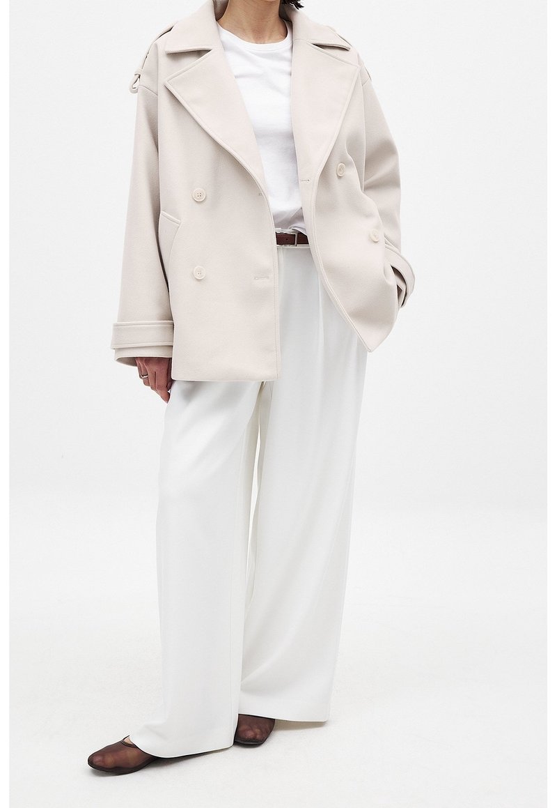 Personne portant un manteau beige croisé sur une chemise blanche et un pantalon blanc large avec des chaussures marron foncé, debout devant un fond uni.
