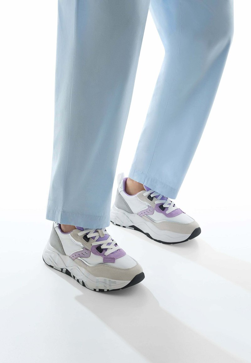 Sneaker bianche e grigie con accenti viola, caratterizzate da una suola spessa, materiali testurizzati e suola in gomma nera. Indossate con pantaloni azzurri chiari.