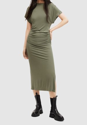 Femme portant une robe midi froncée vert olive associée à des bottes en cuir noir épaisses montant à mi-mollet, debout avec une main sur la hanche.