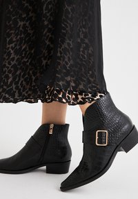 Botines negros con textura de cocodrilo, que presentan un cierre lateral y unacento de hebilla dorada, combinados con una falda de estampado de leopardo en negro y marrón.