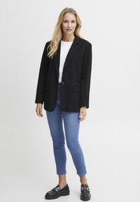 Črni blazer z prilagojenim dizajnom, zapiranje z enim gumbom in dvema sprednjima žepoma, v kombinaciji z modrimi ožjimi kavbojkami in črnimi čevlji.