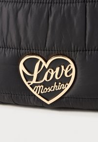 Saco de tecido acolchoado preto com um logotipo dourado em forma de coração, onde está escrito "Love Moschino" em uma elegante caligrafia. Textura suave e acabamento brilhante.