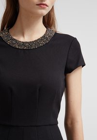 Schwarzes Kleid aus strukturiertem Stoff, mit kurzen Ärmeln und einem mit Metall- und Edelsteinakzenten verzierten Perlenkragen. Taillierte Passform mit Falten.