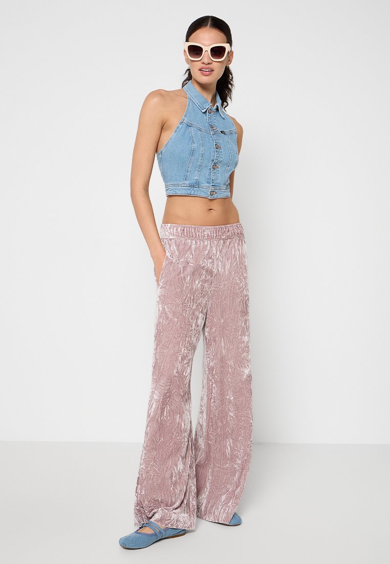 Kobieta w białych okularach przeciwsłonecznych, w bezrękawnikowej niebieskiej dżinsowej bluzce, różowych welurowych szerokich spodniach i niebieskich płaskich butach, stojąca na białym tle.