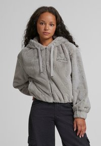 Grå faux fur hættetrøje med lynlås, elastiske manchetter og en afslappet pasform. Har et lille mærkelogo på brystet.