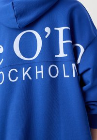 Sudadera azul fabricada con un tejido suave que presenta un texto blanco que dice "O' Stockholm". El diseño incluye capucha y un corte relajado.