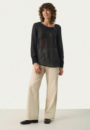 Navy polka-dot blouse, lichtgewicht stof, ronde halslijn met knoopdetail. Crème-brede flanellen broek en zwarte schoenen.