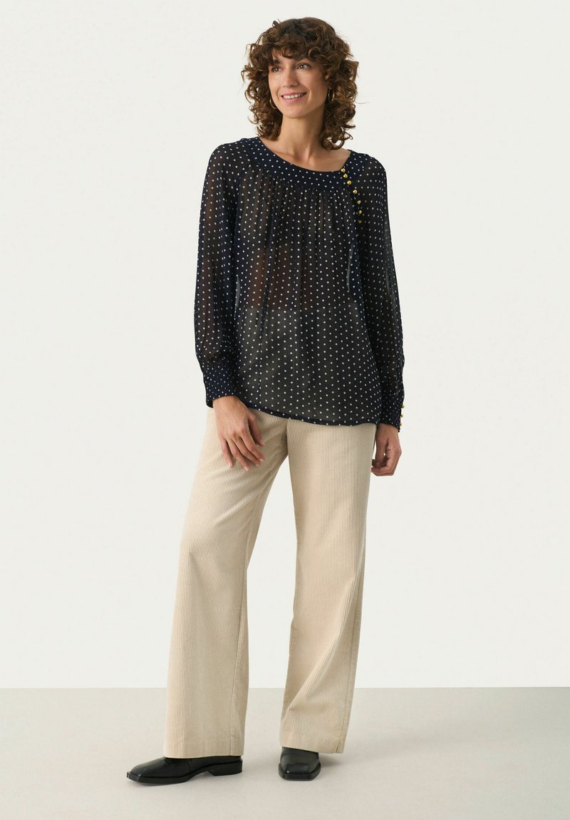 Blouse à pois navy, en tissu léger, col rond avec détail de bouton. Pantalons en velours côtelé crème à jambe large et chaussures noires.