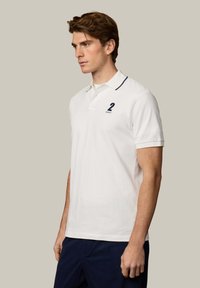 Jeune homme aux cheveux bruns portant un polo blanc à bordure marine avec un logo numéro 2, associé à un pantalon bleu foncé, debout de profil.
