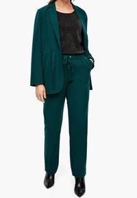 Femme portant un costume vert foncé avec un blazer ample et un pantalon à cordon, associé à un haut noir scintillant et des bottines noires pointues.