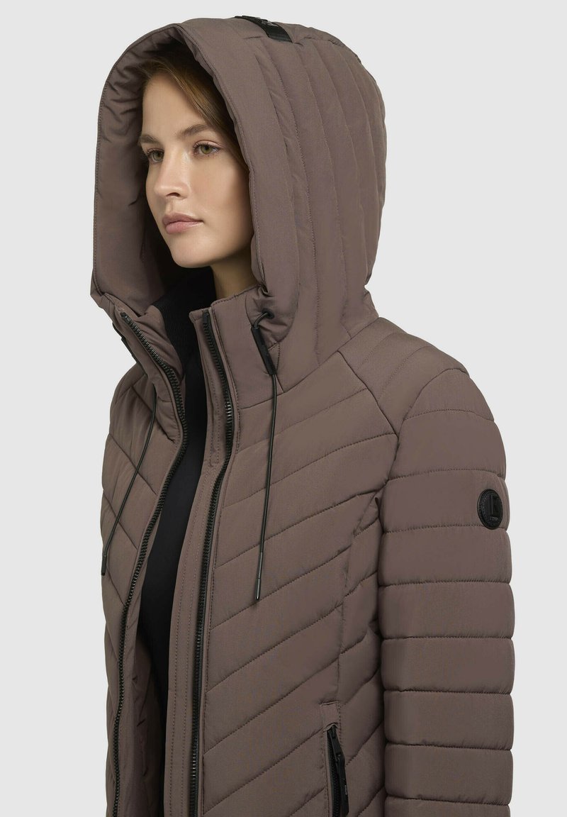 Mujer que lleva una chaqueta acolchada marrón con capucha, con costuras diagonales y horizontales, cremallera negra y cordones ajustables en la capucha.