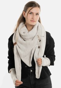 Jonge vrouw met lang haar, die een grote gebreide crèmekleurige sjaal,vingerloze handschoenen, een zwarte jas en donkere jeans draagt, tegen een witte achtergrond.