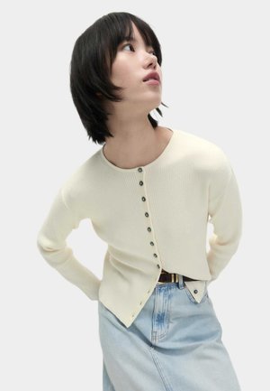 Cream ribbed knit cardigan with round neckline, green buttons, and angled hem. Paired with a light blue denim skirt.