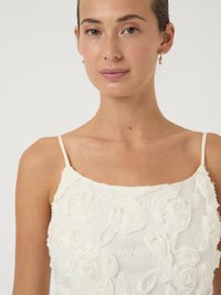 Γυναίκα που φοράει μια λευκή, εμπριμέ, υφασμάτινη, αμάνικη μπλούζα με λεπτές τιράντες, με ελαφρύ μακιγιάζ και μικρές σκουλαρίκια κρίκους σε απλό φόντο.