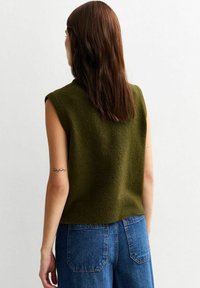 Groene mouwloze gebreide top met een gestructureerde afwerking, gecombineerd met high-waisted blauwe spijkerbroek. Gezien vanuit de achterkant, wat de eenvoud van het ontwerp toont.