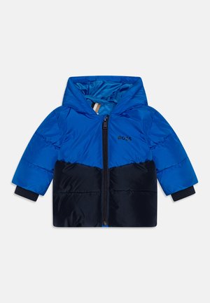 Veste matelassée bleu et marine avec capuche, fermeture éclair sur le devant et détail de logo. Présente des poignets côtelés et deux poches latérales.
