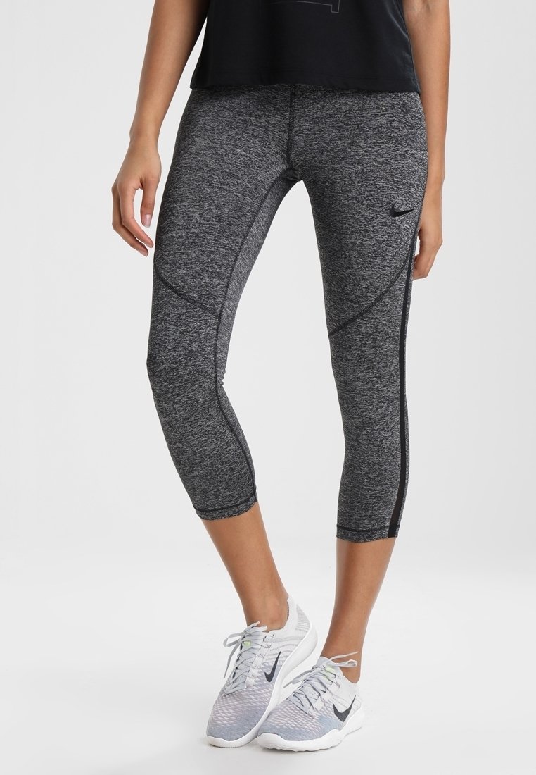 Femme portant des leggings courts gris Nike et des chaussures de running blanches Nike, debout devant un fond uni.