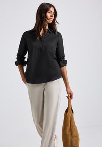 Camicetta nera con maniche arrotolate, abbinata a pantaloni color crema. Tessuto morbido con vestibilità rilassata. Borse in suede in mano.