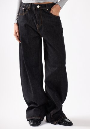 Person iført løstsiddende sorte denimjeans med orange syning og sorte sko, den ene hånd i lommen og den anden hvilende ved siden.