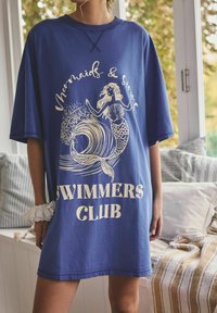 Person trägt ein übergroßes blaues Hemd mit dem Text "Mermaids & Sirens Swimmers Club" und einer Meerjungfrauen-Grafik, steht drinnen in der Nähe einer Fensterbank.